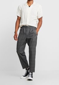 Kortärmad krämfärgad skjorta med ett geometriskt mönster, matchad med mörkgrå rutiga byxor och svarta högskor.