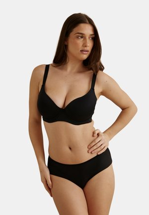 Femme portant un soutien-gorge noir rembourré et un slip noir assorti, debout avec une main sur la hanche, regardant vers sa gauche, sur un fond uni.