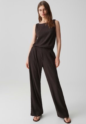 Frau steht, trägt ärmelloses schwarzes strukturiertes Oberteil und passende weite Hosen mit Sandalen, neutraler grauer Hintergrund.