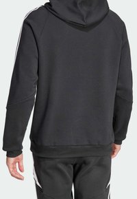 Uomo che indossa una felpa nera con cappuccio e pantaloni da tuta abbinati con strisce bianche sulle maniche e sui lati, visto da dietro.