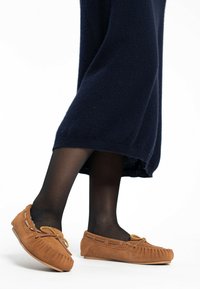 Tan suède moccasin-slippers met stiksels en strikdetail, met een platte rubberen zool, gedragen met zwarte panty’s en een marineblauwe jurk.