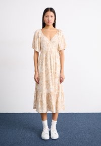 Blomsterprydet, beige omslagskjole med korte, flagrende ermer, samla midje og volangkant. Kombinert med hvite joggesko og sokker.