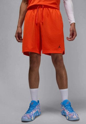 Orange træningsshorts med åndbart mesh-mønster, elastisk talje og snøre. Sort logodetalje på venstre side. Hvide træningssokker og multicolore sneakers fuldender outfittet.