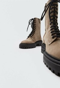 Bottes lacées en suède marron clair avec une semelle noire épaisse en caoutchouc, dotées d'œillets métalliques et d'une surface texturée pour la durabilité et l'adhérence.