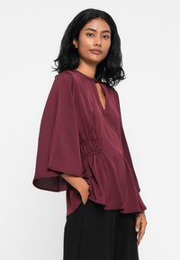 Blusa bordeaux con scollatura a V, maniche ampie e dettaglio arricciato in vita, realizzata in un tessuto morbido e fluido.