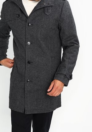 Homme portant un manteau en laine gris foncé boutonné avec une poche poitrine à rabat et des détails de sangles sur le col et les poignets.