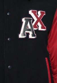 Jachetă din lână neagră cu mâneci din satin roșu, având un logo brodat "AX" în gri, roșu și alb. Închidere cu buton cu snur.