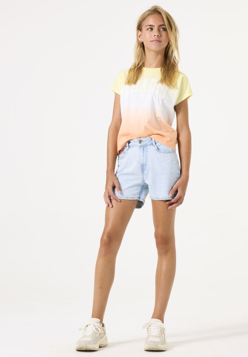 Korte mouwen gradient t-shirt in geel en oranje met 'PARADISE' print; gecombineerd met lichtblauwe denim shorts en beige sneakers.