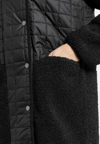 Musta tikattu takki, jossa on teksturoitu, pörröinen alaosa. Varustettu painonapeilla ja sivutaskuilla pehmeästä, lämpimästä kankaasta.