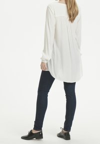 Chemisier blanc à manches longues en tissu texturé, avec un ourlet arrondi, associé à un jean skinny foncé et des chaussures noires à lacets.