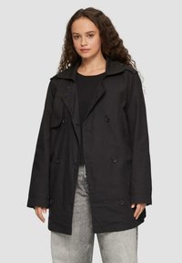 Schwarzer Trenchcoat aus einem weichen Stoff, mit einem doppelt geknöpften Design, großen Revers und Knopfdetails. Getragen über einem schwarzen Oberteil und grauen Jeans.