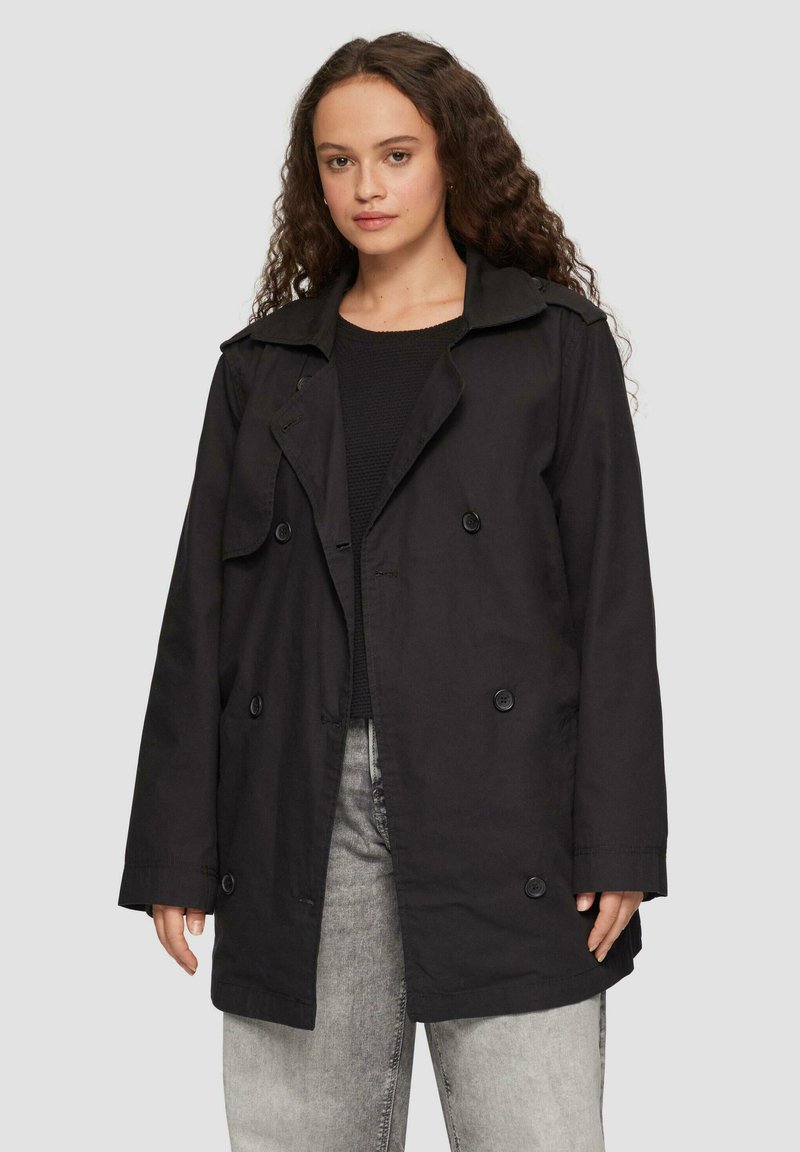 Schwarzer Trenchcoat aus einem weichen Stoff, mit einem doppelt geknöpften Design, großen Revers und Knopfdetails. Getragen über einem schwarzen Oberteil und grauen Jeans.