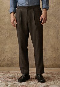 Man wearing dark brown pleated trousers, black leather loafers, and a blue rolled-up long sleeve shirt, standing on patterned rug.