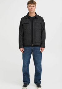 Chaqueta de ante negra con cuello, dos bolsillos en el pecho, cierre con cremallera y mangas largas, llevada sobre una camisa negra y jeans azules.