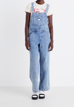 Jasnoniebieskie ogrodniczki jeansowe o luźnym kroju, z przednią kieszenią, regulowanymi ramiączkami i bocznymi kieszeniami, zestawione z kremową koszulą z krótkim rękawem.