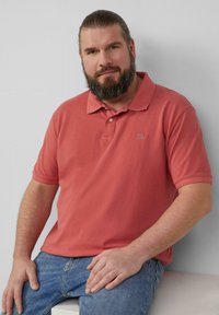 Koralfowa koszulka polo z krótkim rękawem i plisą na dwa guziki, zestawiona z niebieskimi dżinsami. Materiał wydaje się gładki z minimalną teksturą.