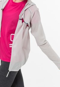 Cortavientos ligero y translúcido de color gris con capucha, detalles reflectantes y puños elásticos, llevado sobre una camiseta rosa que absorbe la humedad y leggings negros.