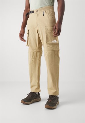 Homme portant un pantalon cargo convertible beige avec poches à rabat à l'avant et ceinture noire, assorti à des chaussures de randonnée foncées sur un fond uni.