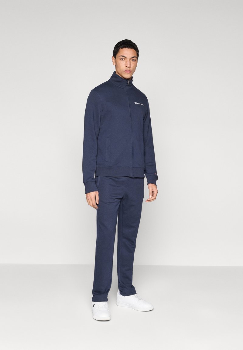 Ζακέτα σε ναυτικό μπλε με φερμουάρ και παντελόνι σετ από βαμβακερό μείγμα υφάσματος· διαθέτουν πλαϊνές τσέπες και μικρό λογότυπο στη ζακέτα.