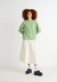 Zelená bavlněná mikina, bílá midi sukně, černé kloboukové boty na podpatku a bílé ponožky; volný střih, měkká textilie a jednoduchý design.