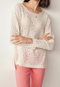 Blouse en dentelle crème avec découpes florales et manches longues, associée à un pantalon rose clair. Caractérisée par un col rond et des fentes latérales à l'ourlet.