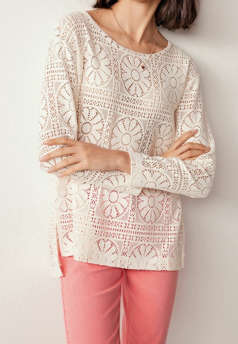 Blouse en dentelle crème avec découpes florales et manches longues, associée à un pantalon rose clair. Caractérisée par un col rond et des fentes latérales à l'ourlet.