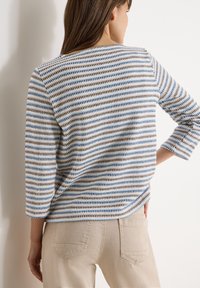 Femme portant un pull en tricot rayé avec des motifs horizontaux bleus, blancs et marron, un pantalon beige, debout contre un mur blanc.