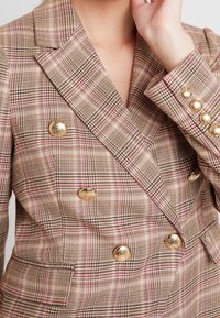 Primo piano di un blazer a scacchi beige e rosa con bottoni decorativi dorati indossato da una persona con una mano alzata vicino al viso.