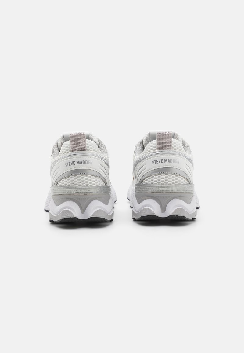 Grijze en witte sport schoenen met een gestructureerde mesh bovenkant, een duidelijke gebogen zool en een zachte hiel tab; branding zichtbaar aan de achterkant.