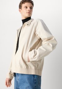 Beige leichte Jacke mit Stehkragen, elastischen Bündchen und Bund, mit einem Reißverschluss und seitlichen Taschen.