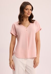 Różowa bluzka z dekoltem w serek i krótkimi, podwiniętymi rękawami, z trzema dekoracyjnymi guzikami. Miękka, gładka tkanina o luźnym kroju. Stylizowana z białymi spodniami.
