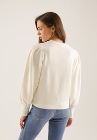 Vrouw met lang bruin haar, gekleed in een witte sweatshirt met pofmouwen en een blauwe spijkerbroek, staand en naar links kijkend tegen een beige achtergrond.