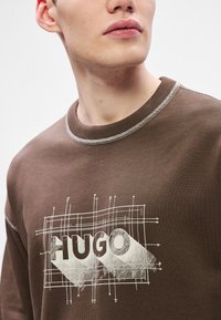 Brun sweatshirt med en grafisk tryck med "HUGO" i stora bokstäver, ljus sömnad och en texturerad, ribbad krage.