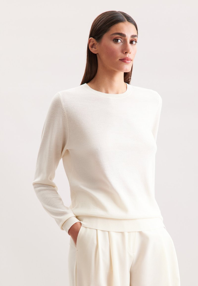 Maglione bianco a maniche lunghe in maglia con scollo rotondo e polsini a coste. Tessuto morbido, vestibilità slim, abbinato a pantaloni chiari.