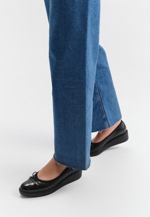 Ballerine in vernice nera con punta arrotondata, dettaglio a fiocco e suola bassa a zeppa, indossate con jeans a gamba larga blu scuro con orlo sfrangiato.