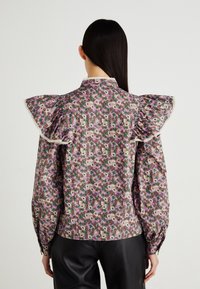 Blomstret bluse med volanger på skuldrene, med et flerfarget mønster på en lys bakgrunn. Kantet med blonder langs kragen og mansjettene.