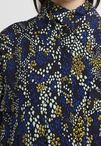 Camicia di tessuto nera con una combinazione di motivi blu, giallo e verde chiaro, dotata di colletto, chiusure a bottoni e due tasche frontali.