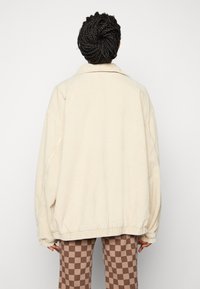 Krémová manšestrová bunda s uvolněným střihem, má límec, spadlé ramena a elastické manžety. Nositá přes hnědé kárované kalhoty.