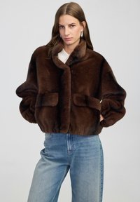 Braune Kunstfelljacke mit hohem Kragen, langen Ärmeln und zwei Vorderkannten. Der Stoff wirkt weich und plüschig und ergänzt lässige blaue Jeans.