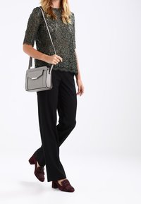 Kobieta ubrana w zielony koronkowy top, czarne spodnie, bordowe obcasy na słupku z ćwiekami oraz nosząca szarą torebkę na ramię.