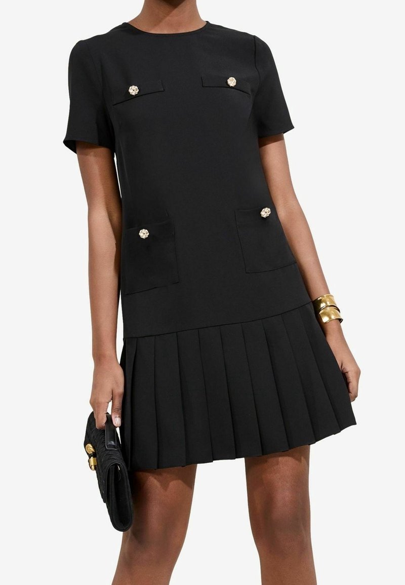 Femme portant une courte robe noire avec jupe plissée, quatre boutons décoratifs sur les poches, tenant une pochette noire et portant des bracelets dorés.