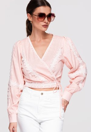 Femme portant de grandes lunettes de soleil marron, un chemisier cache-cœur rose pastel avec un bord en dentelle blanche, et un pantalon blanc taille haute, posant la main sur la hanche.