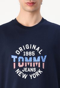 Navy cotton t-shirt featuring a circular graphic with "TOMMY" in a blue and red star-striped pattern, and white text reading "1985 ORIGINAL JEANS NEW YORK."