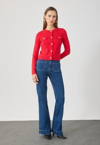 Roter Strick-Cardigan mit Rundhalsausschnitt, Knopfverschluss und zwei vorderen Taschen. Kombiniert mit weit geschnittenen blauen Jeans und schwarzen spitzen High Heels.
