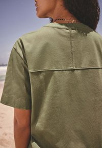Chemise à manches courtes vert olive avec un empiècement, texture douce et coutures visibles, portée devant un décor de plage.