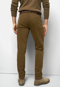 Mann in braunen Slim-Fit-Hosen, beigen Wildleder-Chelsea-Boots, einem braunen Pullover und einem mehrfarbigen gewebten Gürtel, der an einer weißen Wand steht.