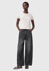 Top rosa chiaro a maniche corte abbinato a jeans scuri a gamba larga con cuciture evidenti e tasche frontali. La modella è in piedi con le mani nelle tasche.