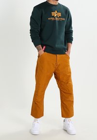 Sudadera verde oscura con el logo "ALPHA INDUSTRIES", combinada con pantalones cortos naranjas que tienen paneles en las rodillas y zapatillas blancas.