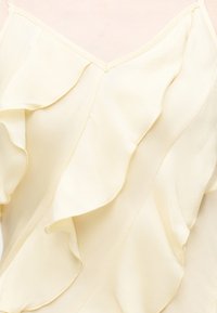 Φόρεμα κρεμ χρώματος με λεπτές τιράντες και κυματιστές λεπτομέρειες βολάν μπροστά και στα πλάγια σε ανοιχτό φόντο.