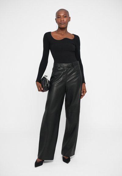 Top negro ajustado de manga larga con escote redondeado, combinado con pantalones de piel negros de pierna ancha. La modelo sostiene un pequeño bolso negro.